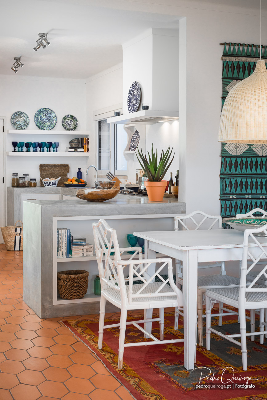 Cozinha luminosa com azulejos turquesa geométricos, ilha em betão, pratos decorativos e mesa branca com cadeiras