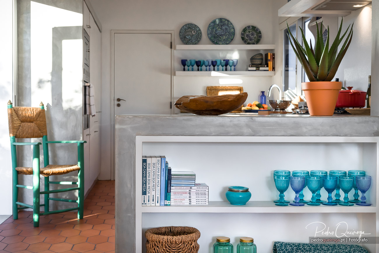Cozinha luminosa com bancada em cimento, pratos decorativos azuis na parede, copos turquesa e cadeira verde vintage