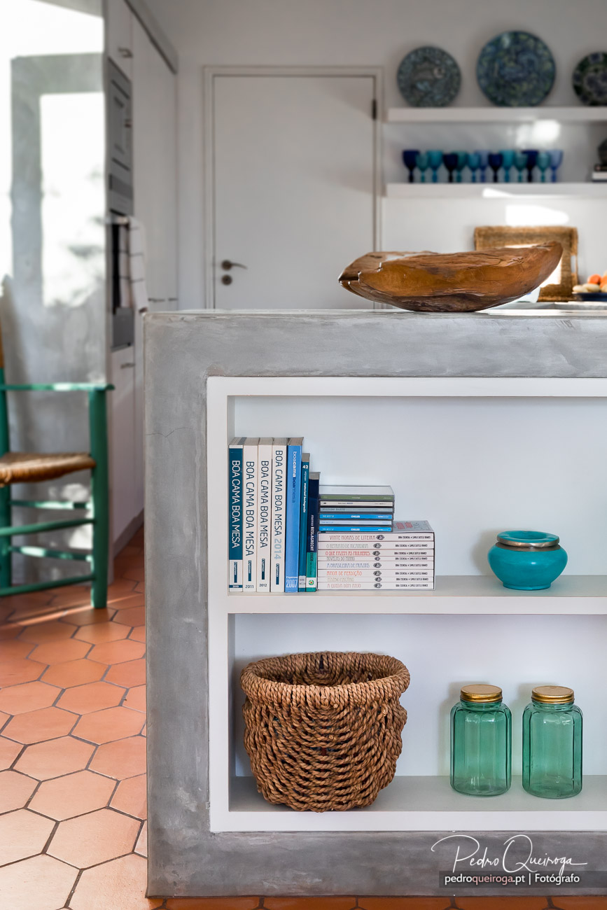 Cozinha moderna com ilha em betão, prateleiras com livros fotográficos e detalhes decorativos em tons azuis e turquesa