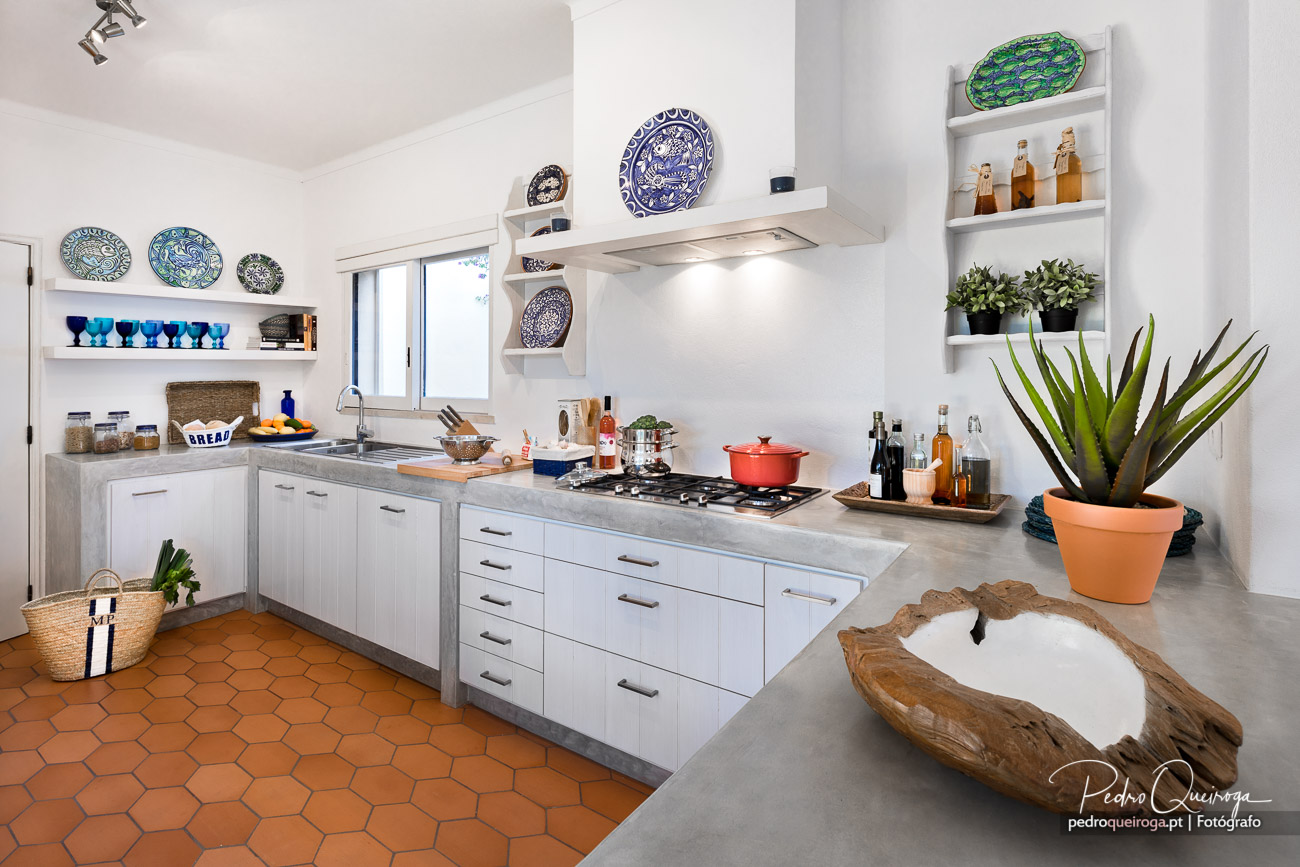 Cozinha moderna com mobiliário branco, bancada em cimento, pratos decorativos, pavimento terracota e plantas ornamentais.