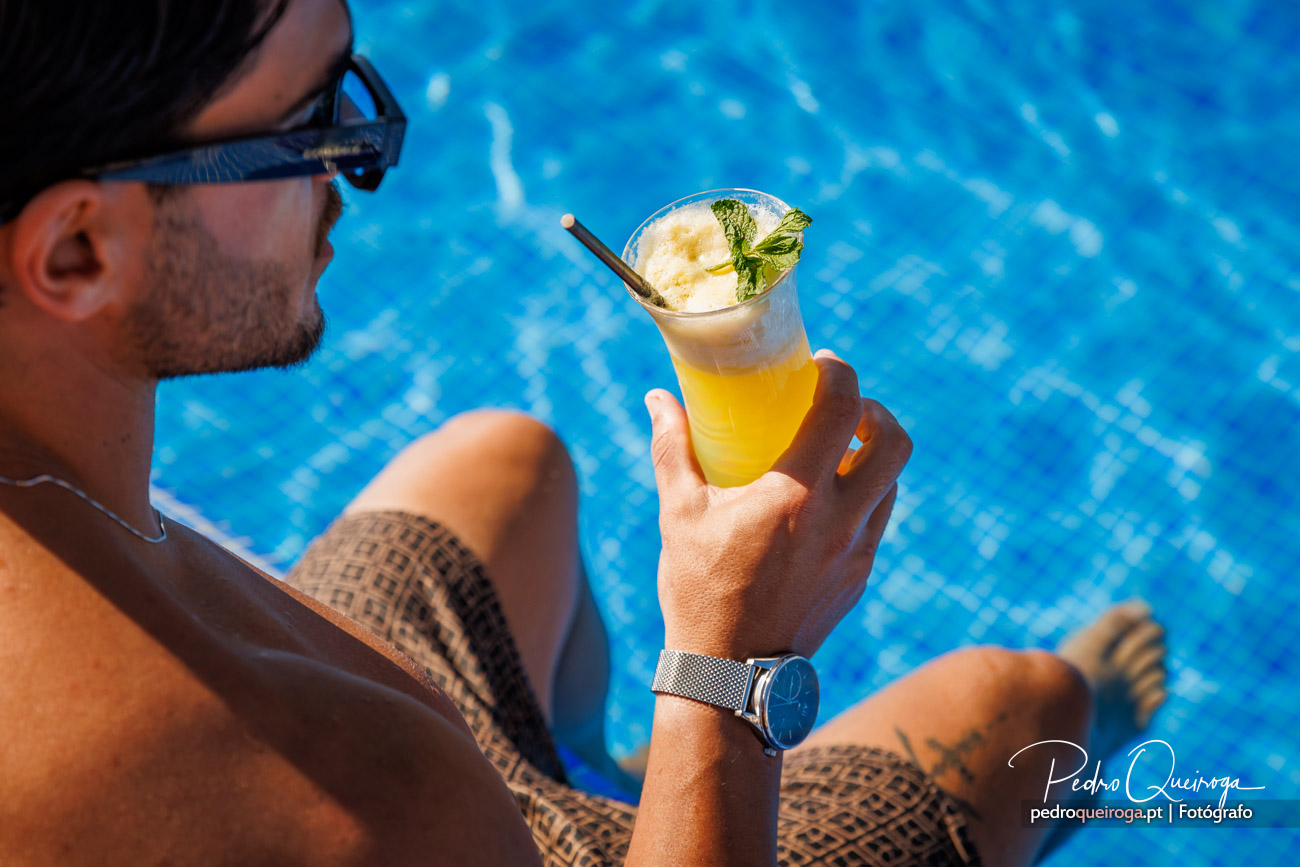 Homem com óculos de sol segura cocktail amarelo decorado com hortelã junto à piscina num dia de verão