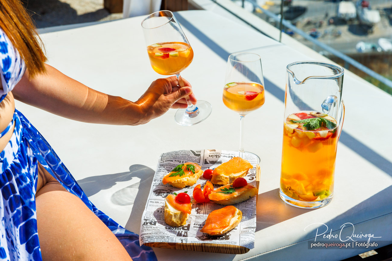 Mão segura copo de sangria ao lado de petiscos e jarra, numa mesa branca com decoração estival e descontraída