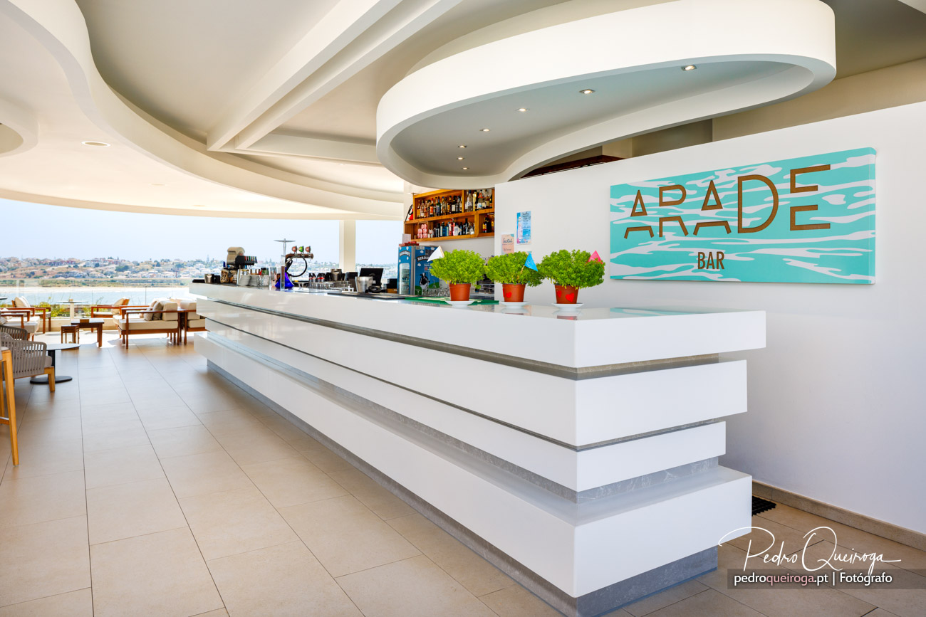 Bar moderno com balcão branco, teto curvo, plantas decorativas e vista para o mar ao fundo num ambiente luminoso