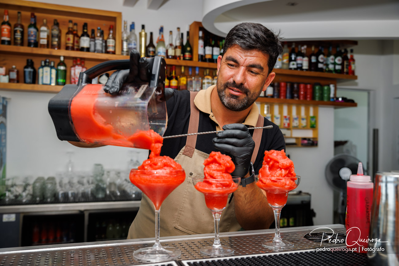 Barman concentrado a servir cocktail vermelho gelado em três copos junto ao balcão de bar repleto de garrafas