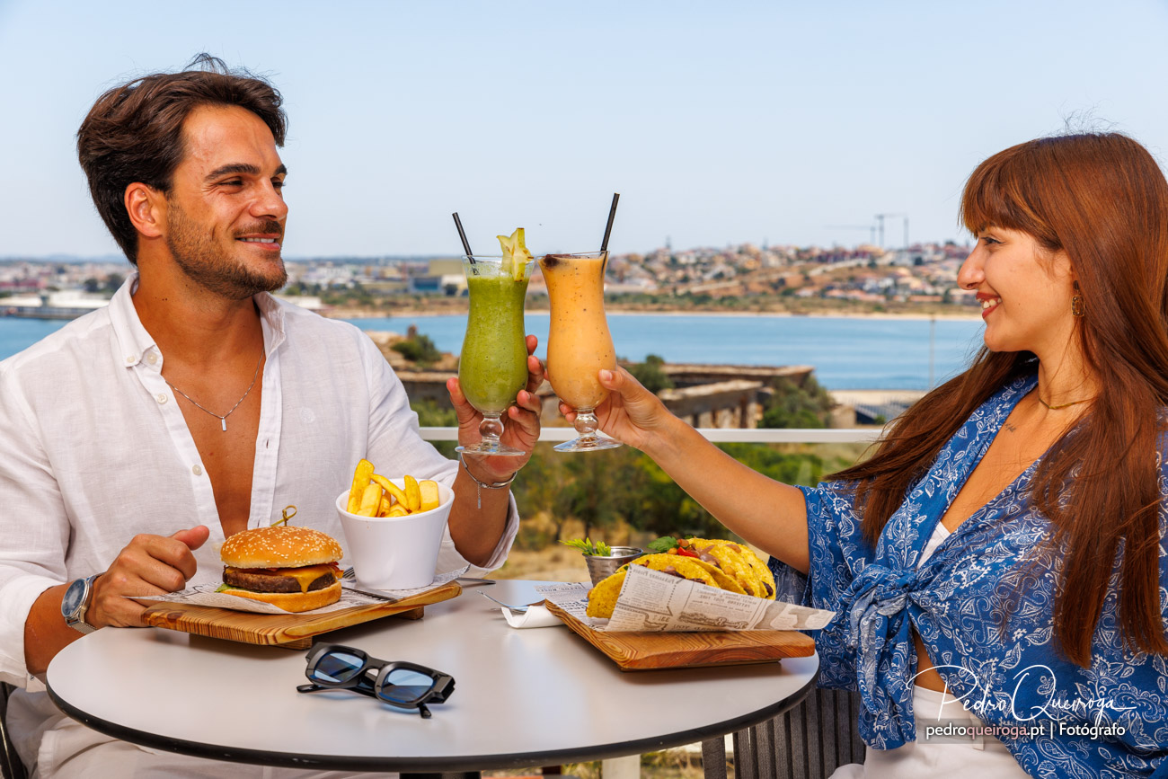 Casal brinda com cocktails coloridos num terraço com vista panorâmica sobre o rio e a cidade ao fundo.