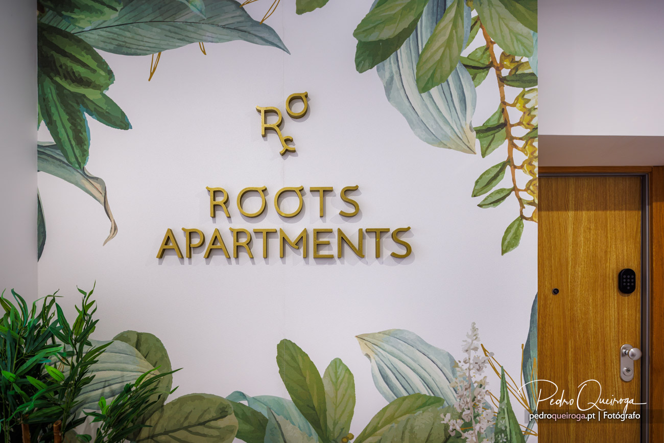 Parede branca com letras douradas Roots Apartments, rodeada por folhagens tropicais pintadas e porta de madeira ao lado