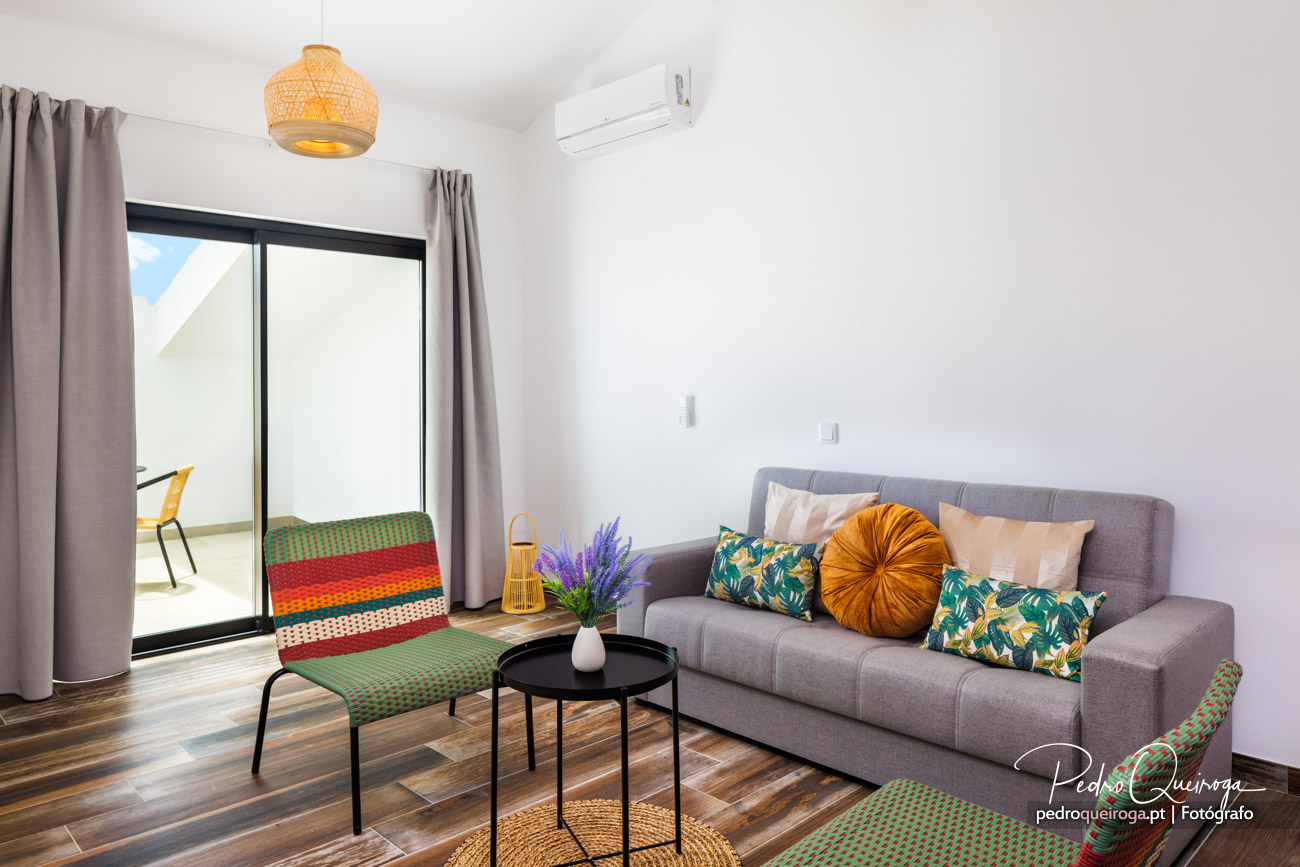 Sala de estar luminosa com sofá cinzento, almofadas coloridas e acesso a terraço com tapete vibrante.