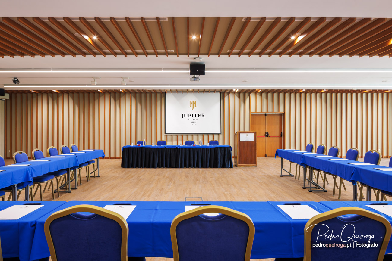 Sala de conferências com mesas em U cobertas por toalhas azuis, ecrã de projeção e teto com ripas de madeira