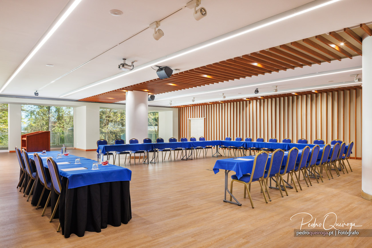 Sala de conferências ampla com mesas em toalha azul, cadeiras arrumadas, teto em ripado de madeira e grandes janelas.