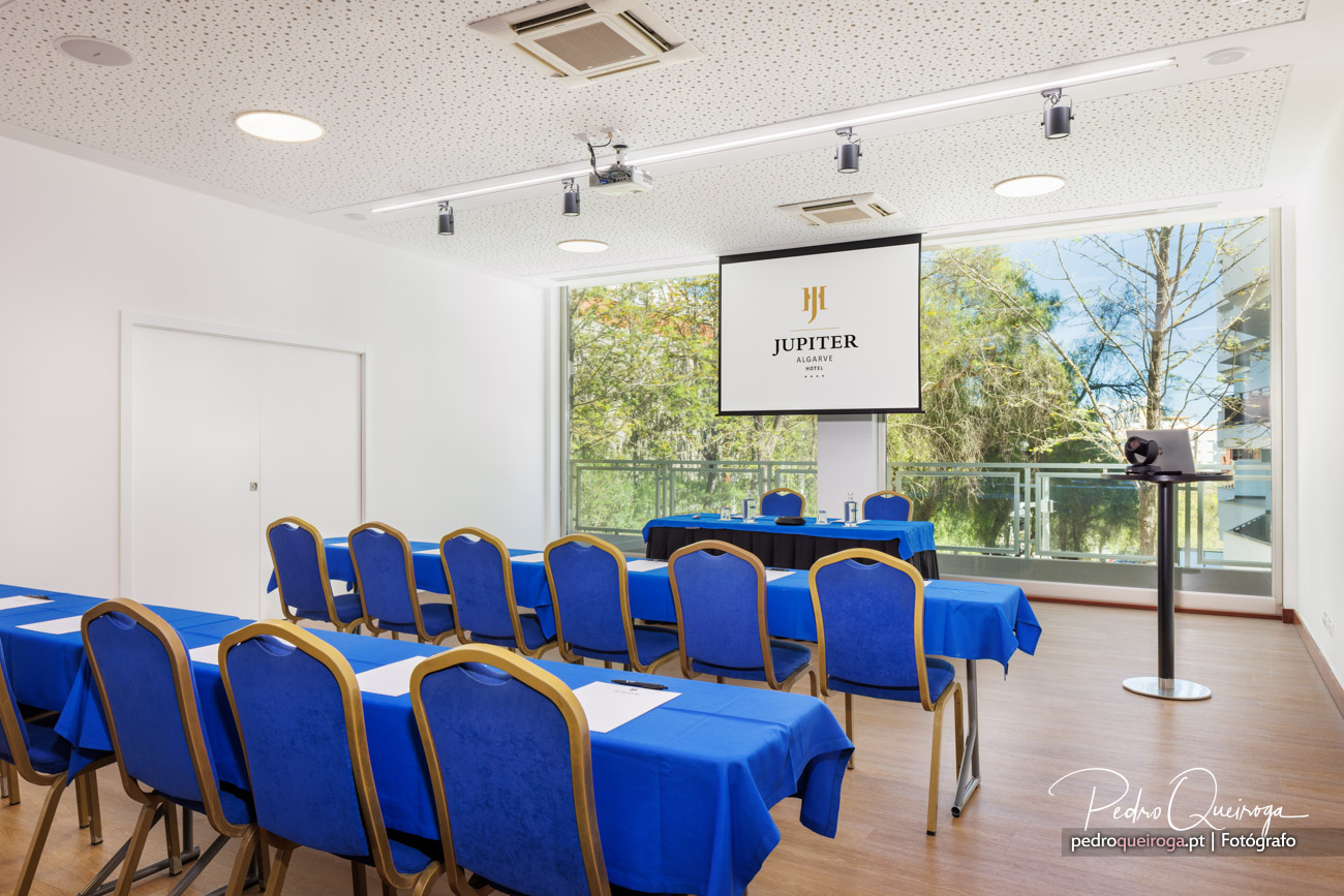 Sala de reuniões com mesas cobertas por toalhas azuis, cadeiras elegantes e ecrã de projeção com logótipo Jupiter Algarve Hotel