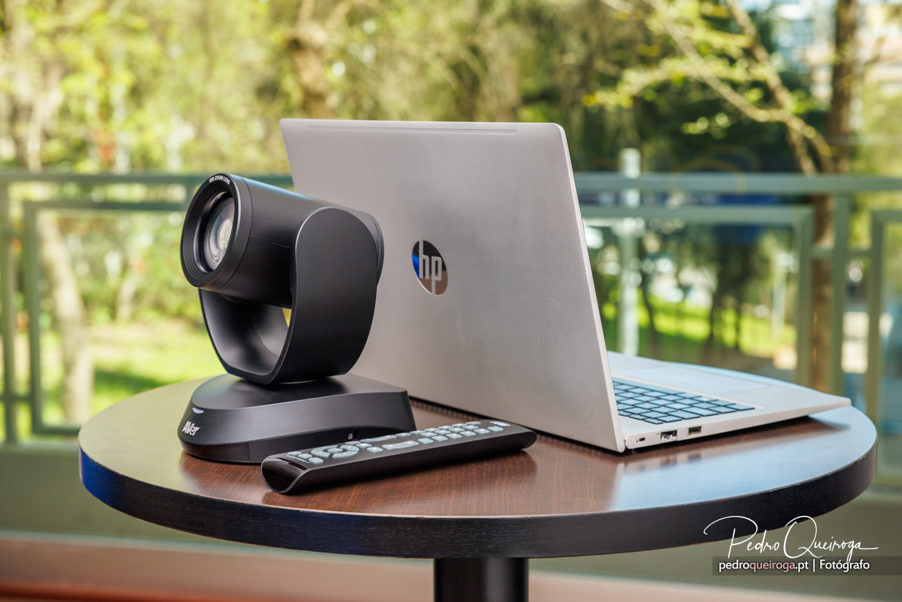 Câmara de videoconferência e portátil HP sobre mesa redonda, com comando e ambiente exterior desfocado ao fundo