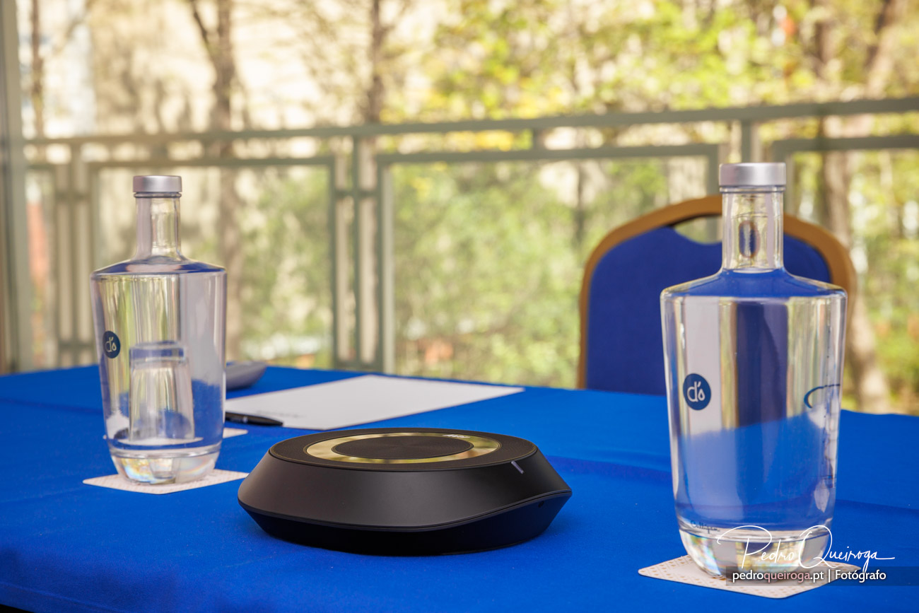 Duas garrafas de água transparentes e dispositivo eletrónico preto sobre mesa azul com cadeira e jardim ao fundo