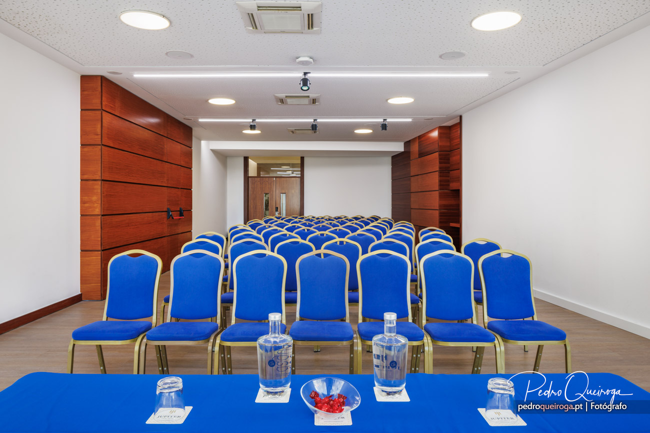 Sala de conferências com cadeiras azuis dispostas em filas, painéis de madeira nas paredes e mesa com garrafas de água.