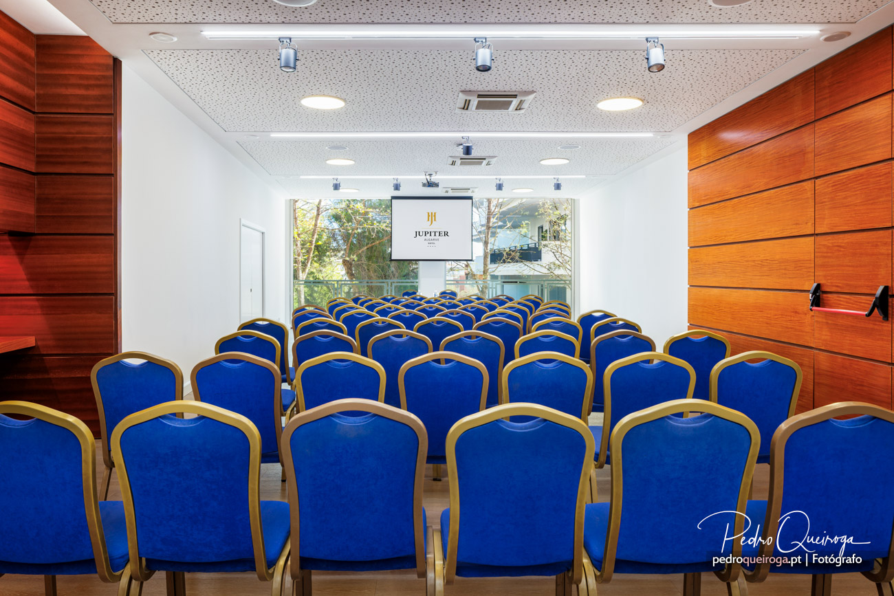 Sala de conferências com cadeiras azuis e douradas, ecrã de projeção ao centro e painéis de madeira nas paredes laterais.