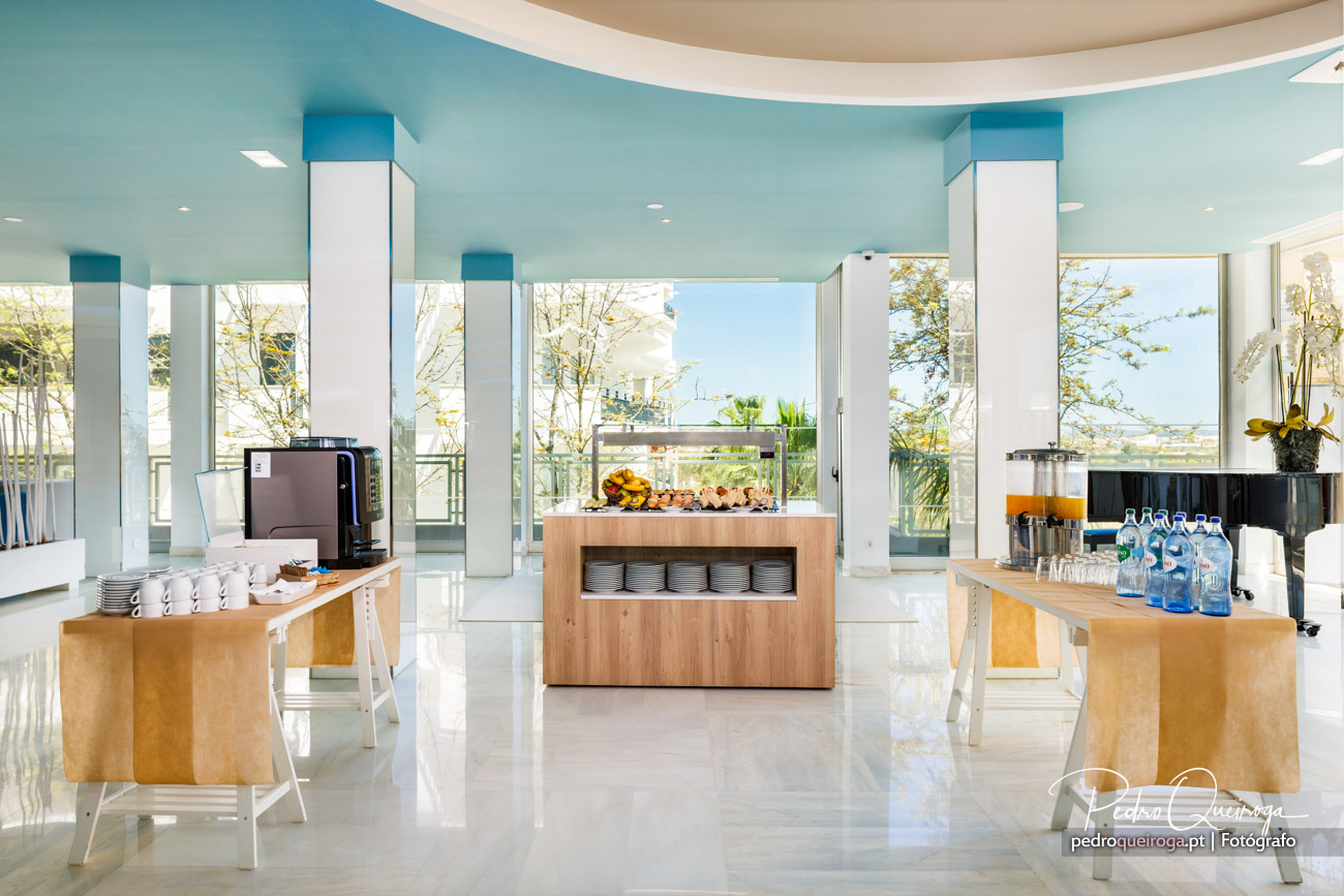 Sala de pequenos-almoços moderna com tetos azuis, colunas brancas, buffet em madeira e amplas janelas panorâmicas.