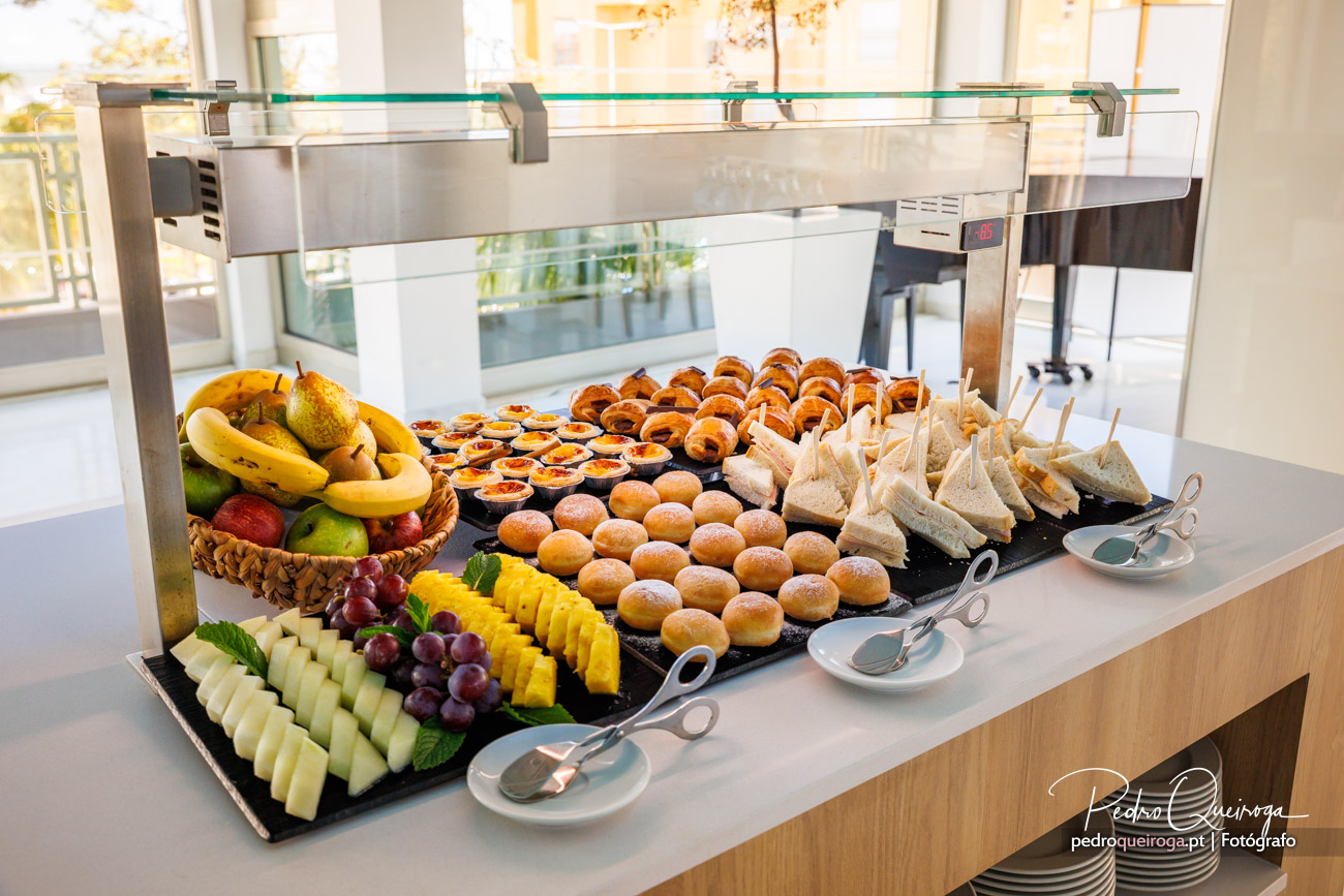 Buffet de pequeno-almoço com fruta fresca, croissants, bolos e sandes num balcão luminoso com vista exterior.