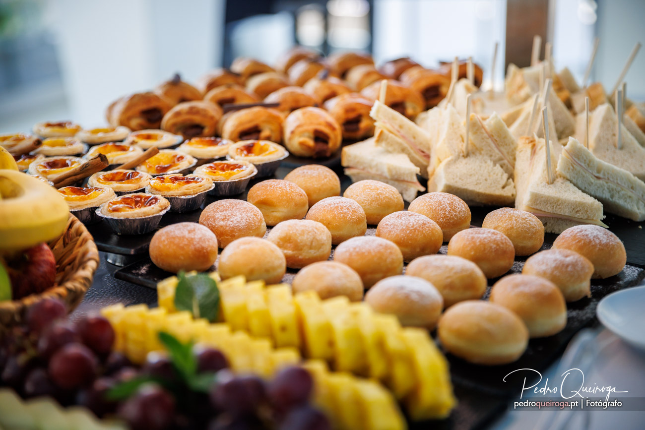 Mesa de buffet repleta de doçaria variada, pastéis de nata, bolas de Berlim, sandes e frutas frescas num evento