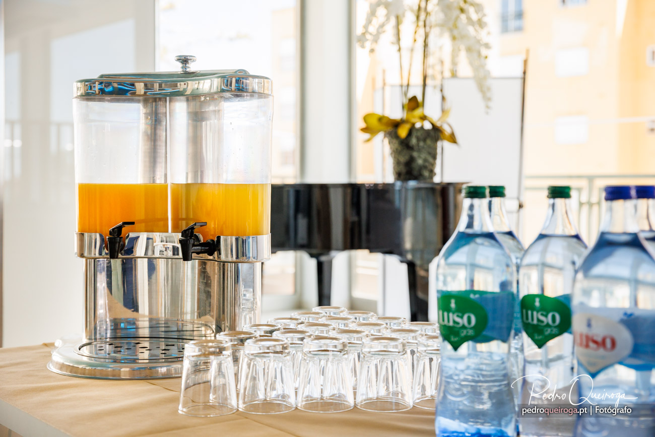 Dispensador duplo de sumo laranja e ananás, copos e garrafas de água, numa composição fotográfica de evento.