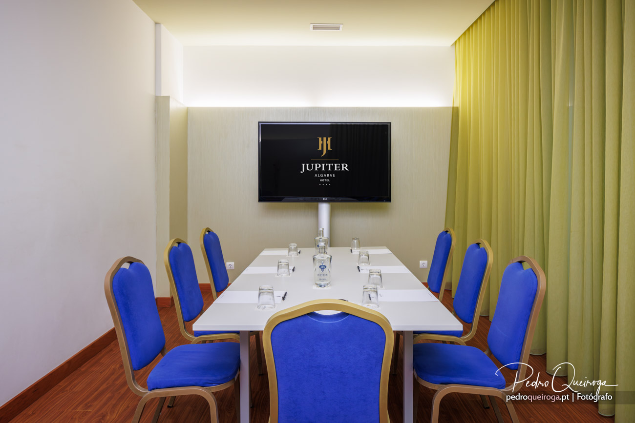 Sala de reuniões elegante com cadeiras azuis, mesa branca, ecrã na parede e cortinas amarelas laterais.