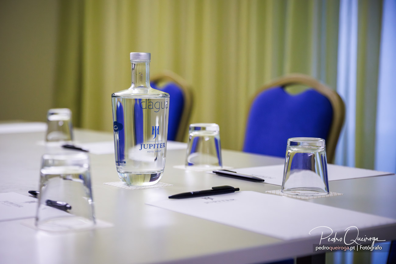 Composição fotográfica de mesa de reunião com garrafa de água Jupiter, copos e cadeiras azuis ao fundo
