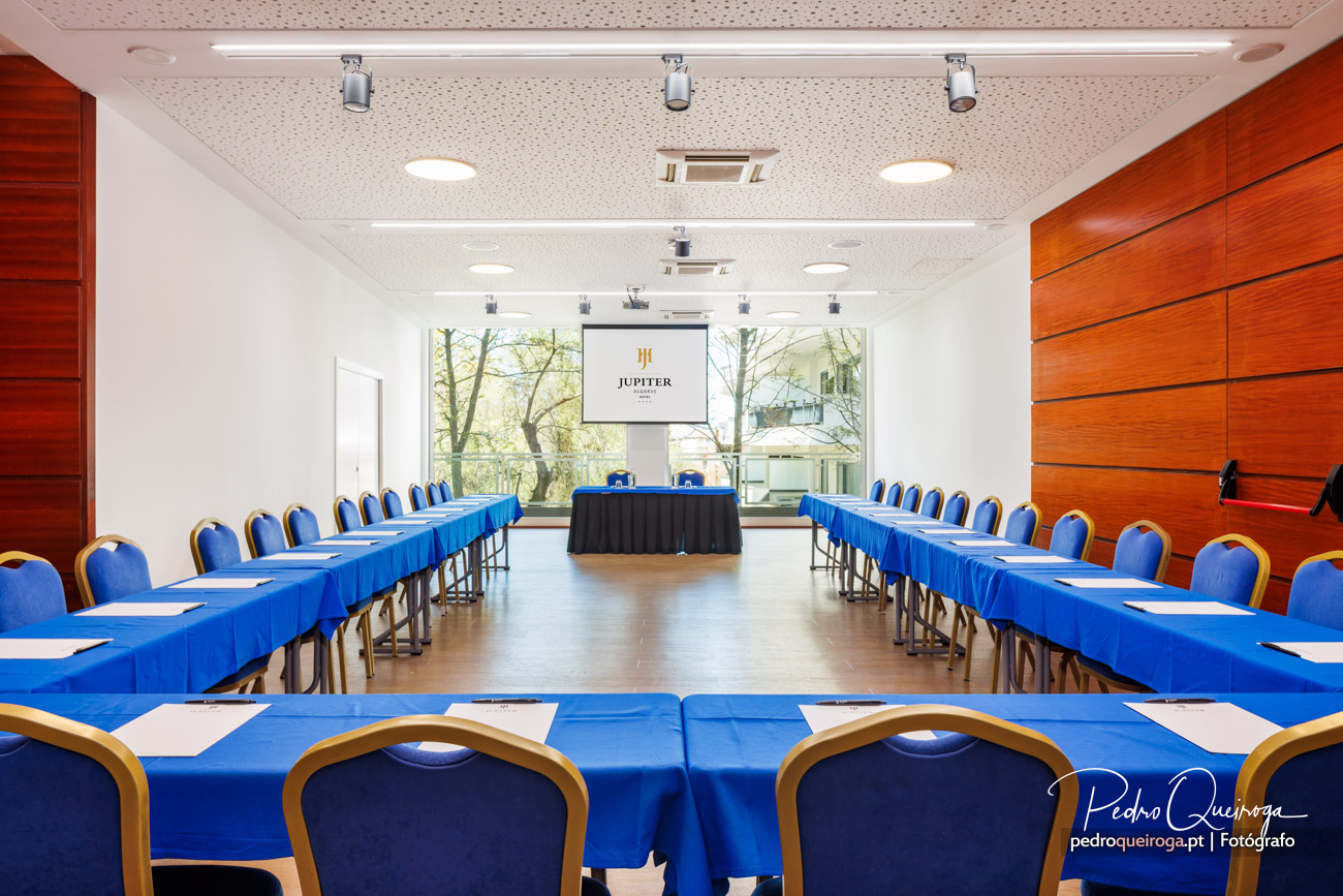 Sala de reuniões em formato U com mesas azuis, cadeiras estofadas e ecrã de projeção ao fundo junto a janelas amplas