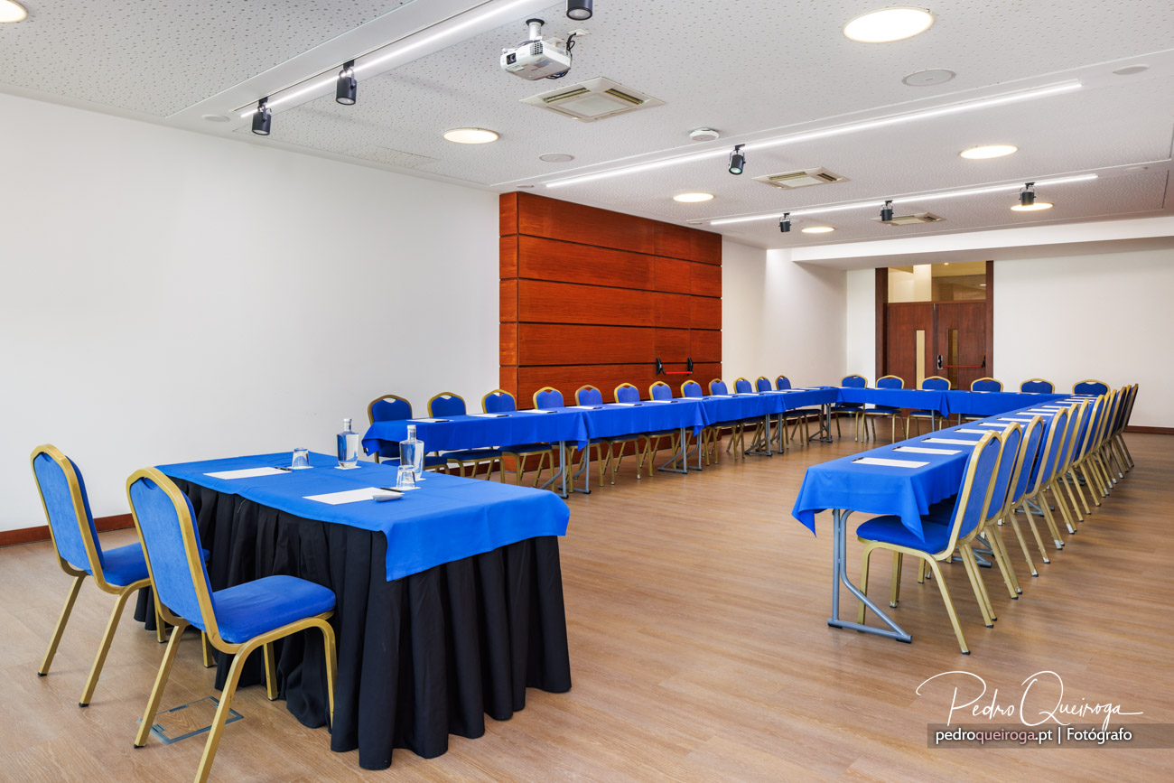 Sala de conferências ampla com mesas em U cobertas por toalhas azuis, cadeiras estofadas e painel de madeira ao fundo