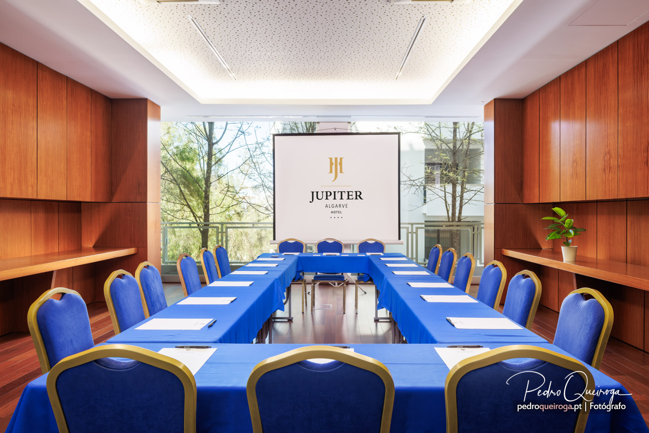 Sala de reuniões elegante com mesas em U forradas a azul, cadeiras estofadas e ecrã de projeção ao fundo.