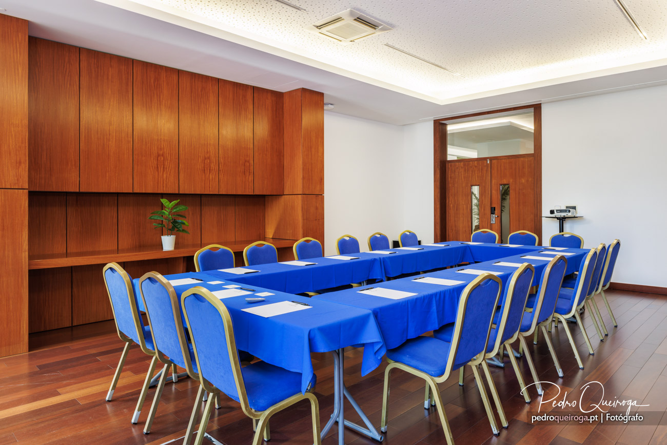 Sala de reuniões com mesas em U cobertas por toalhas azuis, cadeiras estofadas e painéis de madeira na parede