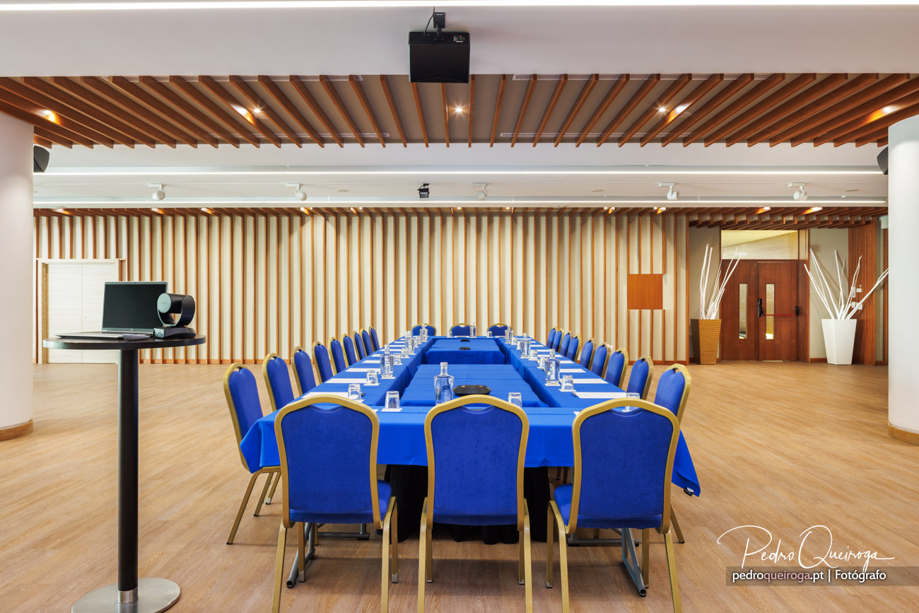 Sala de conferências ampla com mesa em formato U revestida a azul royal, cadeiras estofadas e teto em ripado de madeira
