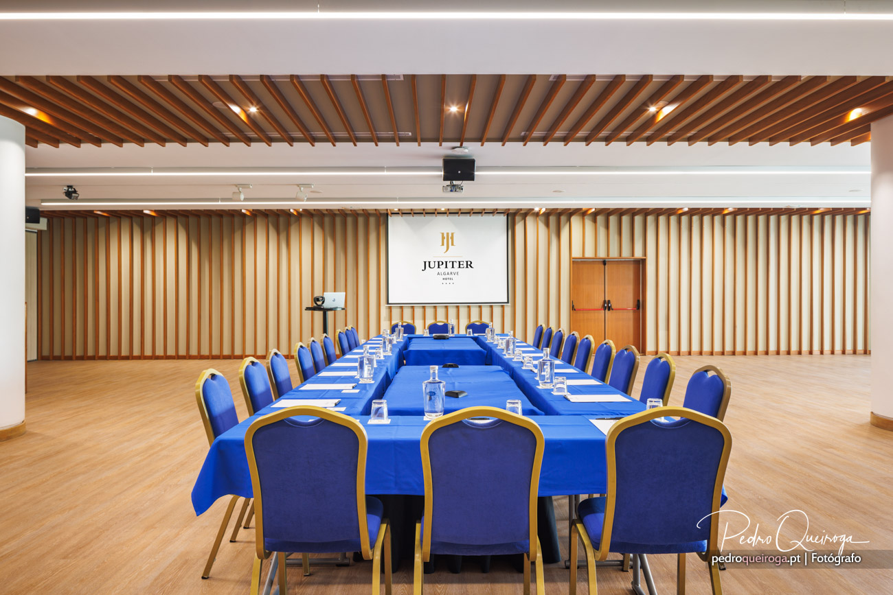 Sala de reuniões com mesa em U coberta por toalha azul, cadeiras estofadas e parede em ripado de madeira com projetor
