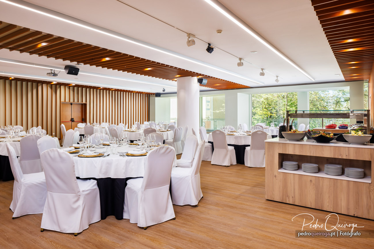 Salão de eventos elegante com mesas postas, teto em madeira ripada, iluminação LED e buffet junto à janela ampla
