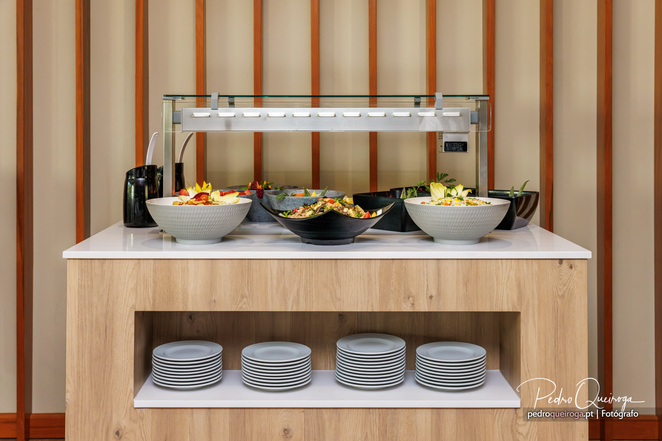 Buffet de hotel com saladas coloridas em tigelas brancas e pretas, decoração em madeira clara ao fundo