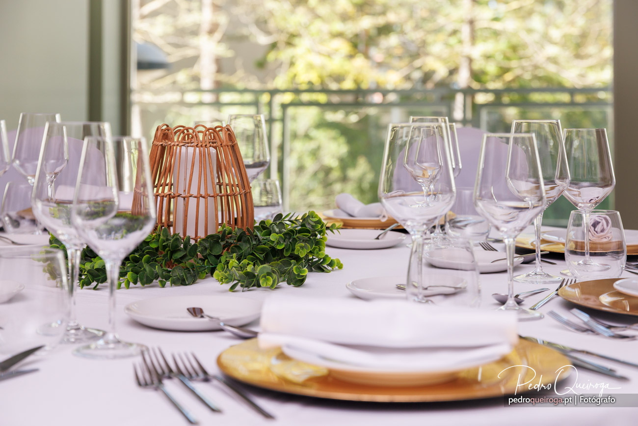 Mesa elegante com toalha branca, copos de cristal, pratos dourados, lanterna de vime e folhagem verde fresca ao centro