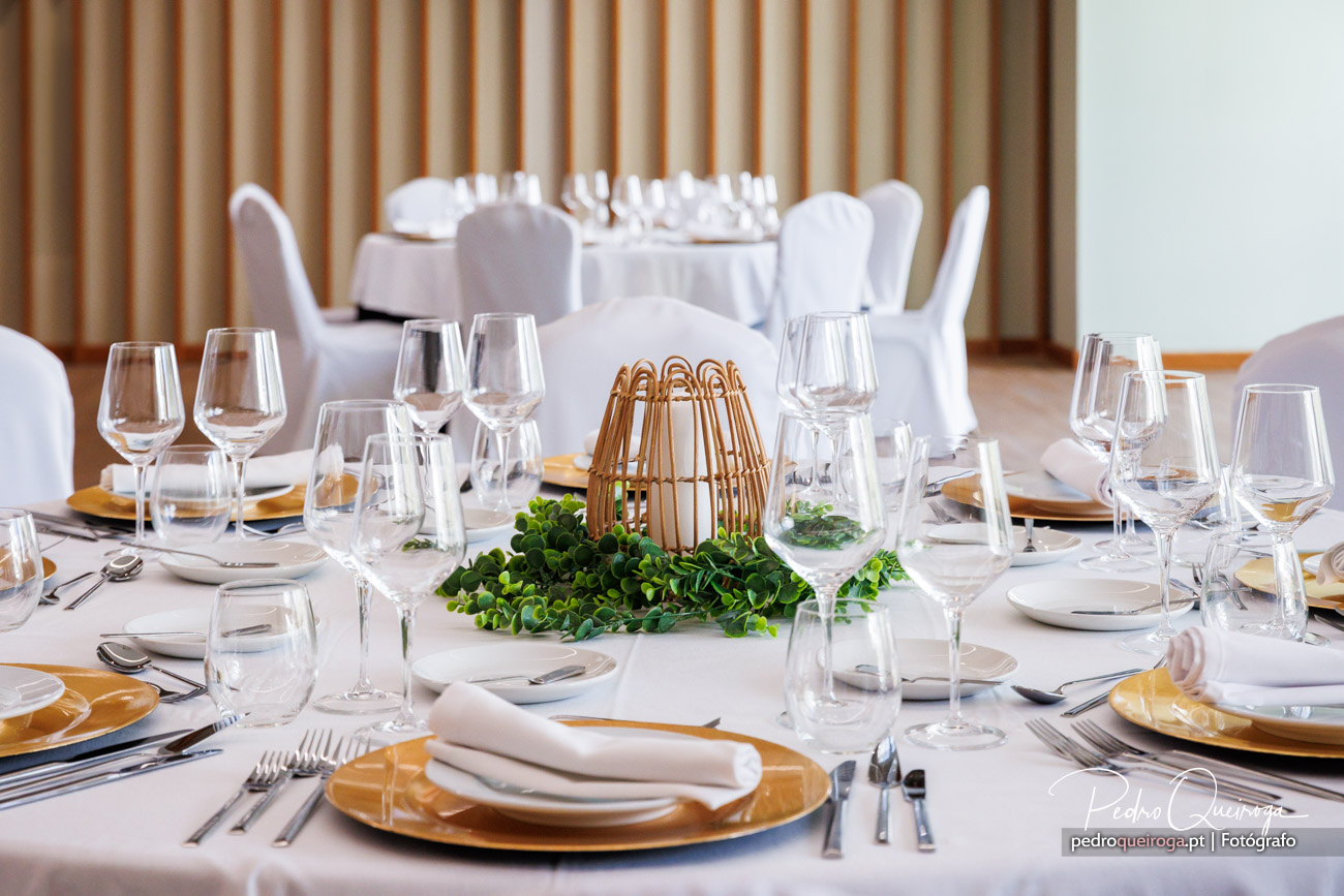 Mesa de banquete elegante com toalha branca, pratos dourados, copos de cristal e centro decorativo em vime com folhagem verde