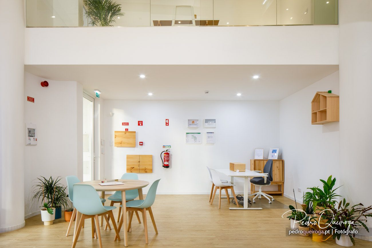 Escritório moderno com mesas, cadeiras turquesa e brancas, mobiliário em madeira, plantas e sinalização de segurança.