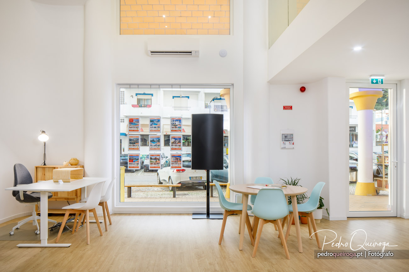 Interior moderno e luminoso com mesas, cadeiras azuis claras, parede branca, janela ampla e detalhes em madeira clara.