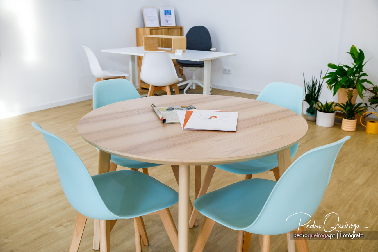 Mesa redonda em madeira clara rodeada por cadeiras turquesa num escritório luminoso com plantas e secretária ao fundo