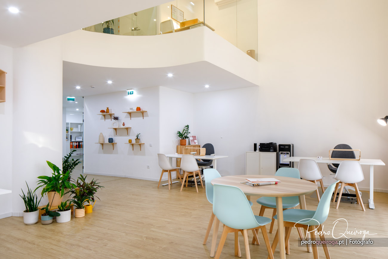 Espaço de trabalho amplo e luminoso com mesas redondas, cadeiras em tons pastel e prateleiras decorativas na parede branca.