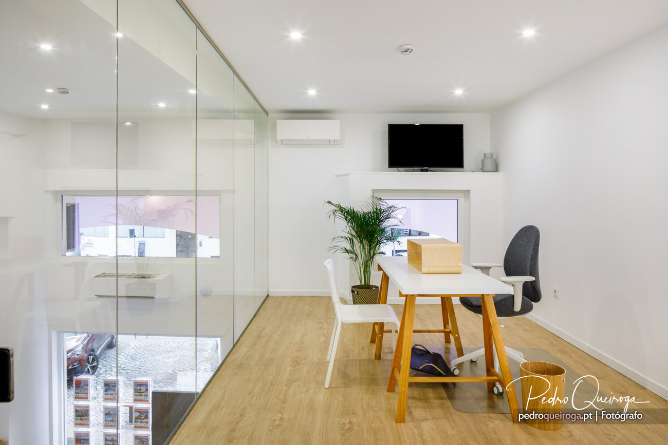 Escritório moderno com parede envidraçada, mesa branca, cadeiras, televisão na parede e iluminação embutida no teto.