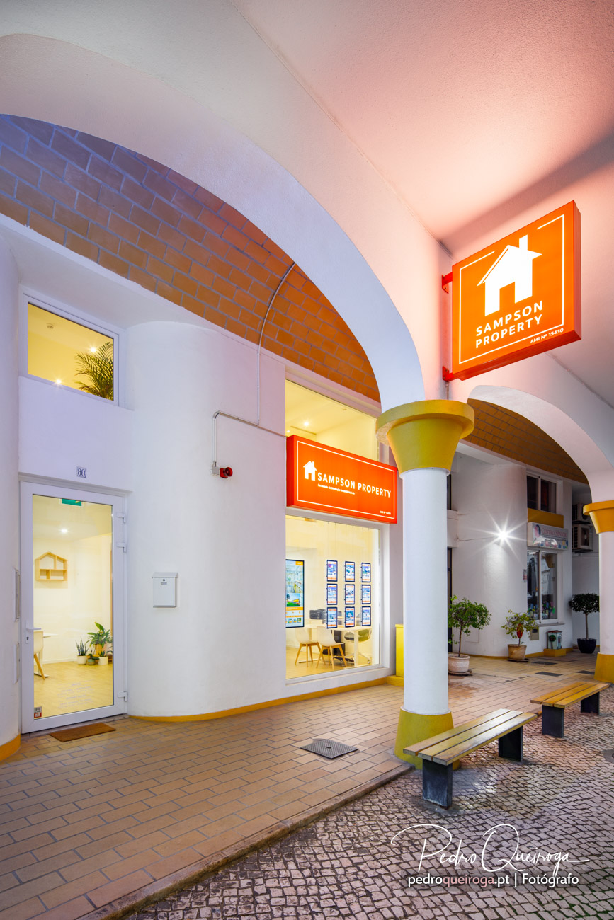 Fachada noturna de agência imobiliária com arcos em tijolo, colunas brancas e amarelas e montra iluminada