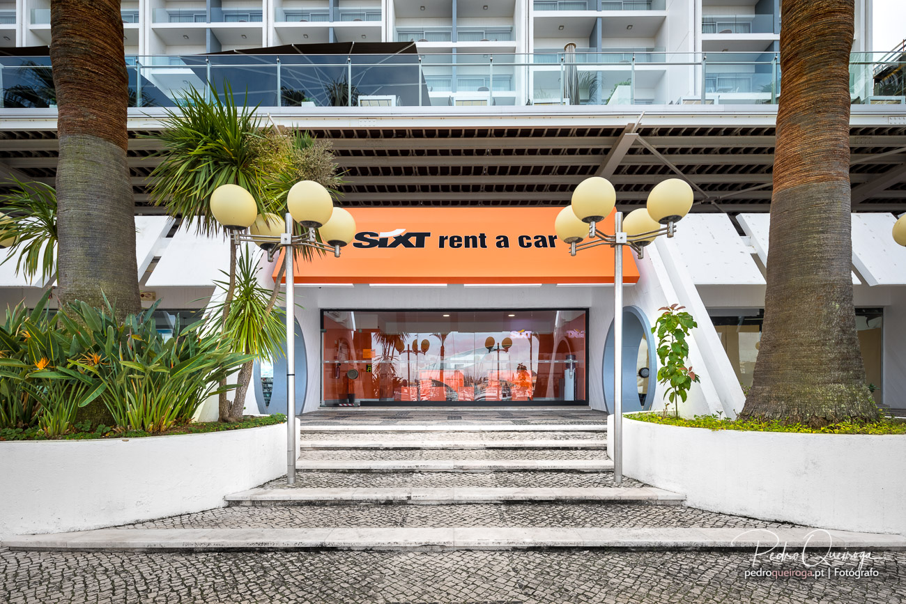 Fachada moderna da Sixt rent a car com palmeiras, candeeiros esféricos e edifício branco ao fundo