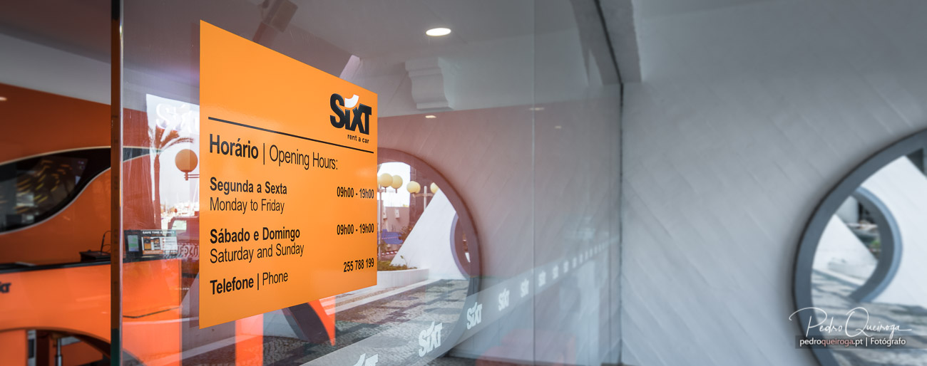 Vidro com horário de funcionamento da Sixt colado, refletindo o interior moderno e laranja da loja de aluguer de carros.