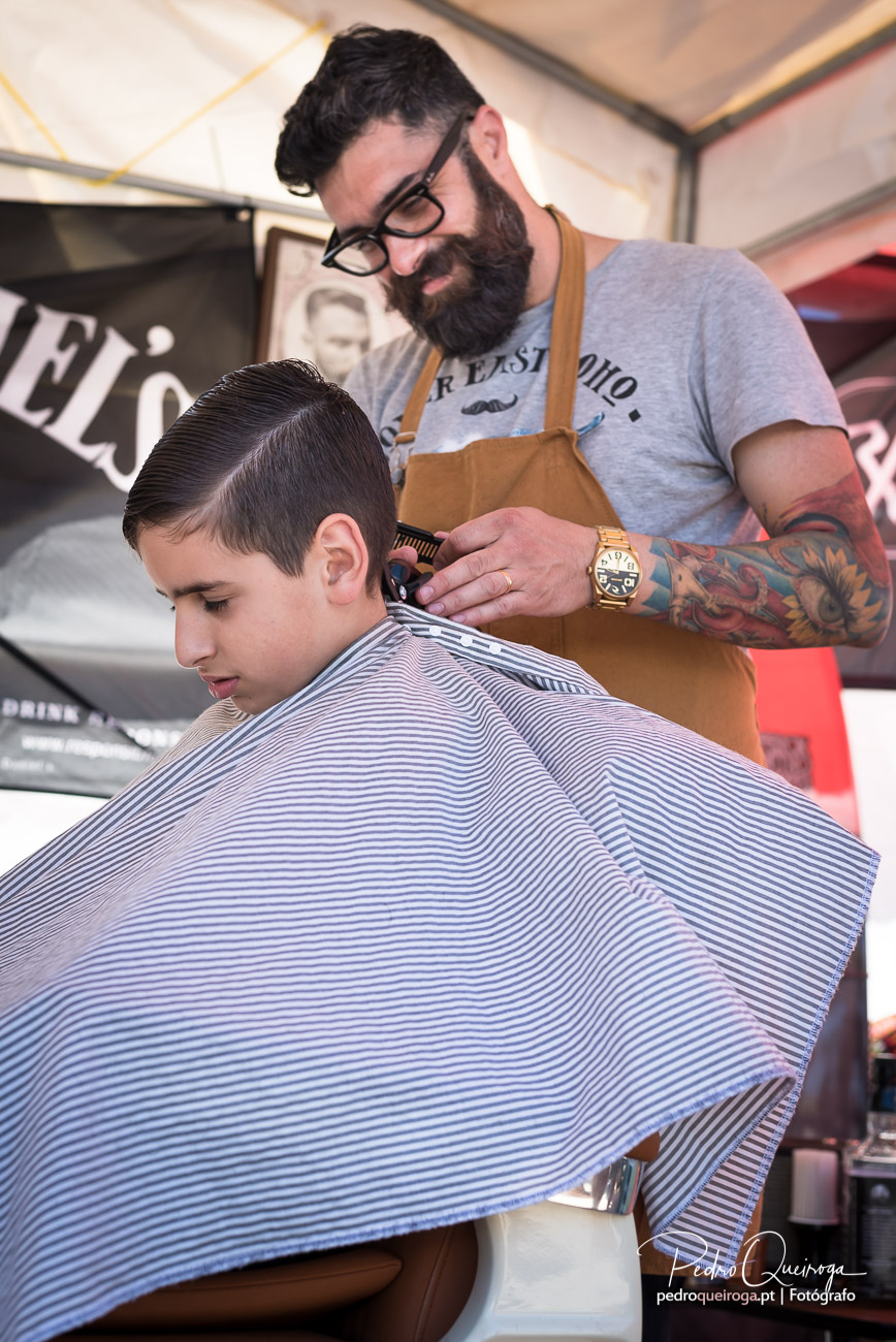 Barbeiro tatuado com avental castanho corta cabelo de jovem cliente coberto com capa às riscas azuis e brancas