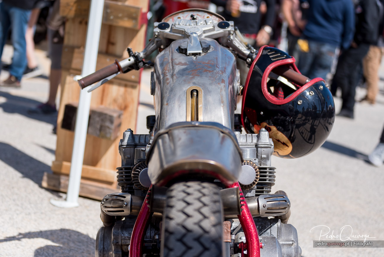 Mota clássica com depósito metálico envelhecido e capacete preto pendurado, imóvel numa exposição ao ar livre