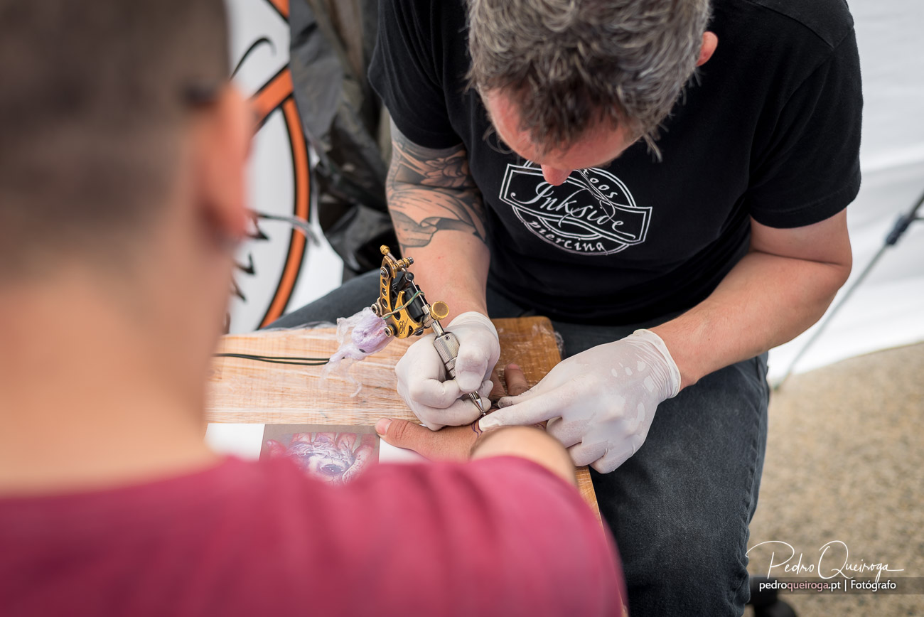 Tatuador concentrado a trabalhar numa tatuagem no braço de cliente com máquina dourada e luvas brancas