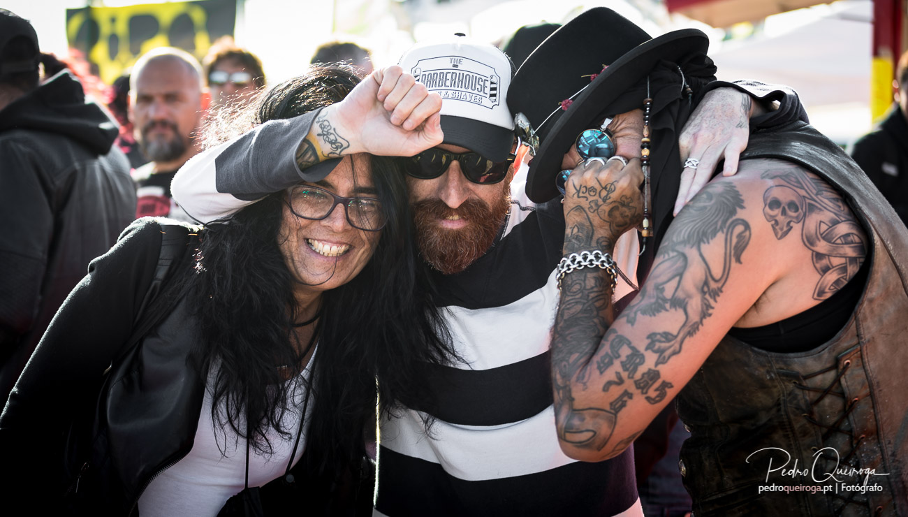 Grupo de amigos tatuados abraçados e sorridentes num evento ao ar livre com ambiente descontraído e animado