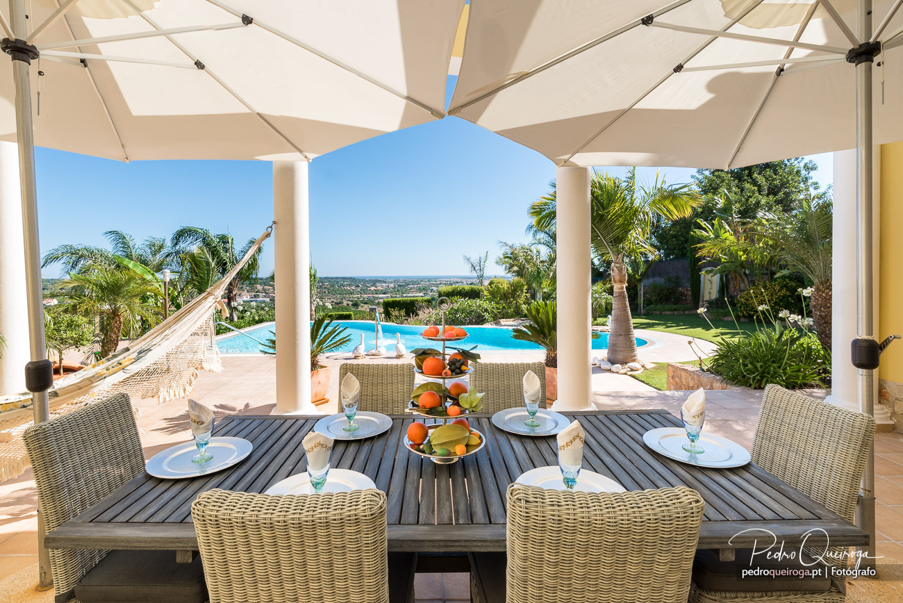 Terraço coberto com mesa posta, rede suspensa, piscina azul ao fundo e jardim tropical sob céu limpo