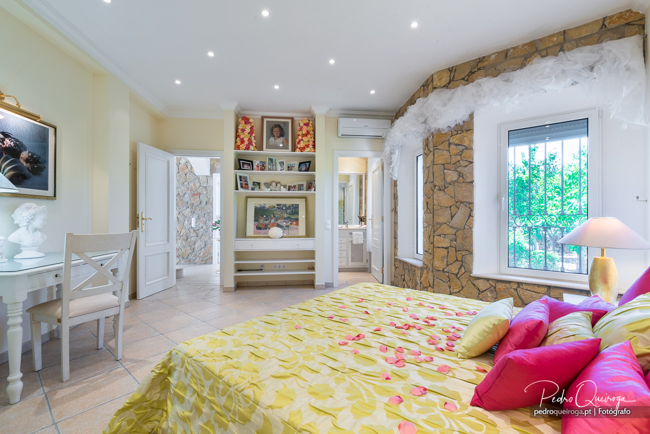 Quarto espaçoso com parede em pedra, cama floral amarela, mesa branca e janela ampla com luz natural abundante.