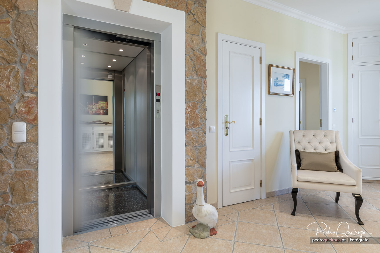 Hall elegante com elevador moderno, parede em pedra natural, portas brancas e poltrona capitoné bege.